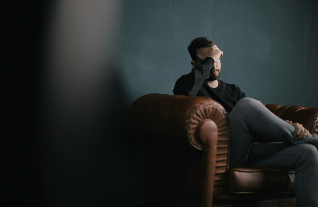 A stressed or distressed man sits on a leather armchair, holding his hand to his forehead, in a dimly lit room with a dark blue wall.