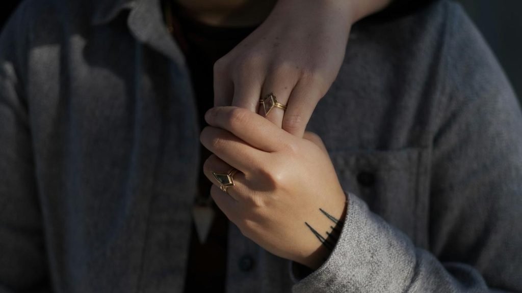 A close-up of two hands gently clasped together, both wearing matching gold geometric rings.