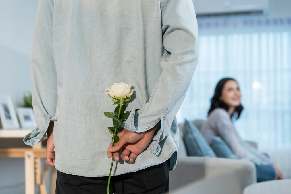 A man holding a white rose behind his back, with a smiling woman sitting on a couch in the blurred background.
