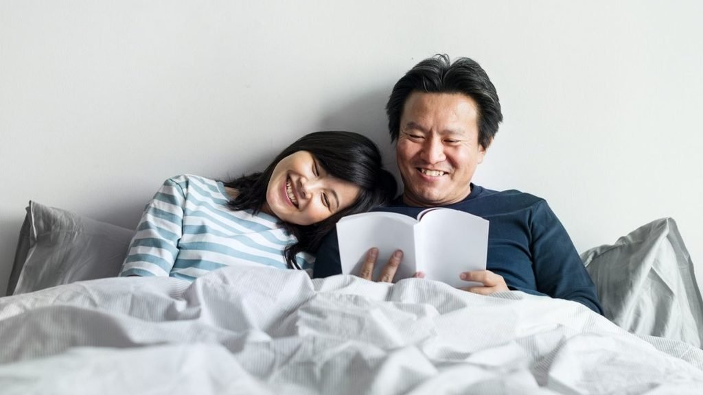 A couple reading a book together in bed with a white wall in the background, both of them smiling.