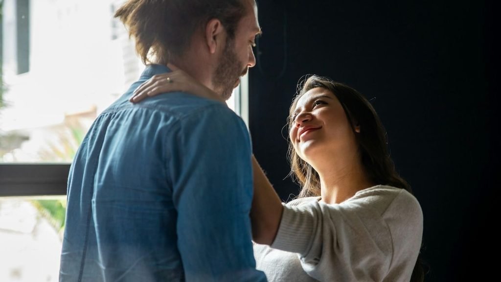 A man in a blue shirt is standing in front of a woman in a white top, who is looking up and smiling at him.