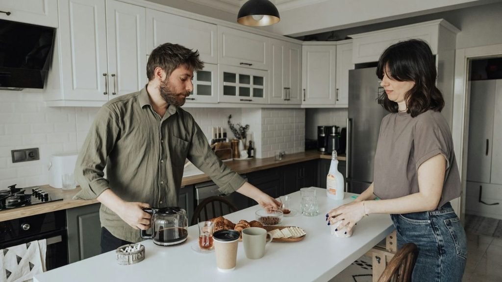 Man pouring coffee and woman standing by white kitchen island with food.
