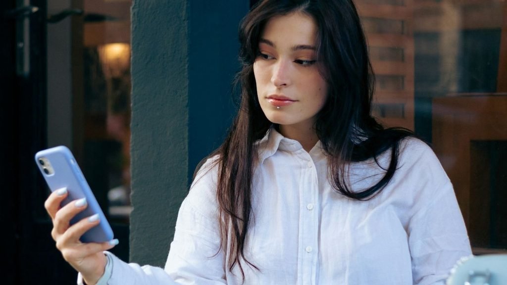 A woman with dark hair and a white shirt is holding a light blue smartphone outdoors, gazing at it.