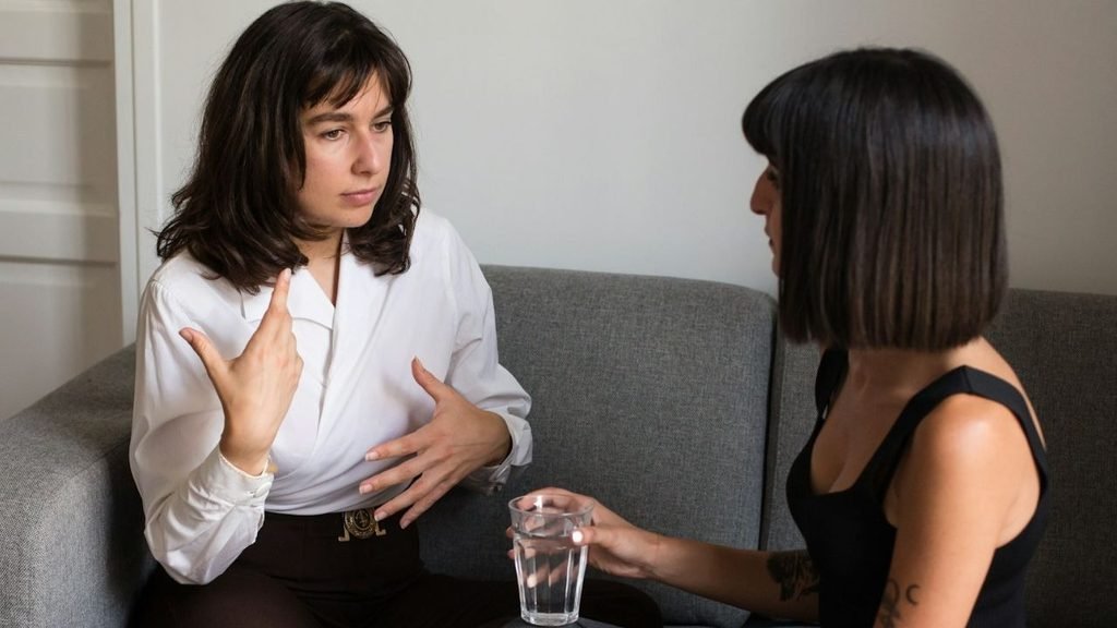 Two women sitting on a couch, one gesticulating with her hands, the other holding a glass of water.