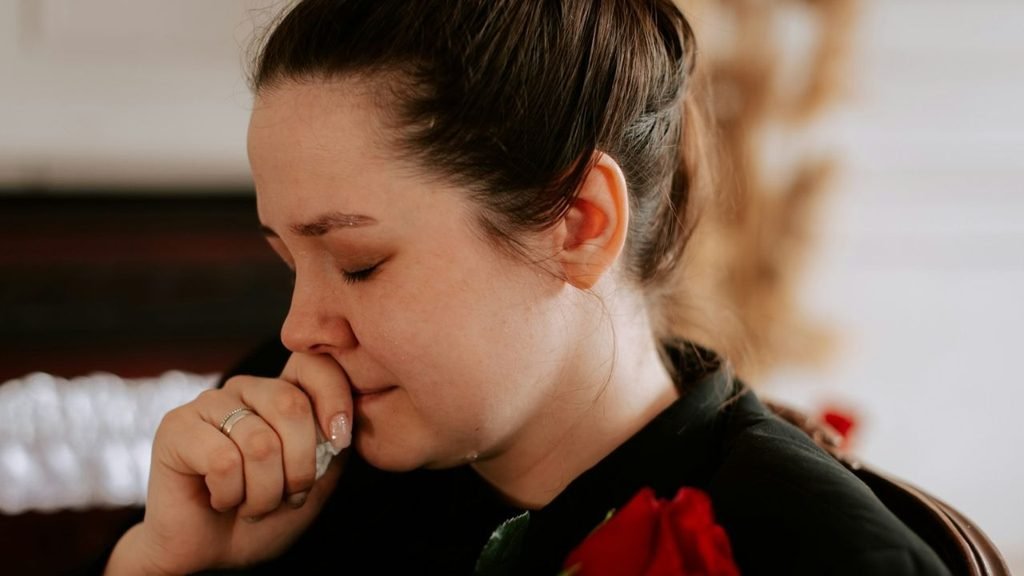 A woman with her eyes closed, holding a tissue to her face, wearing a black top and a ring.
