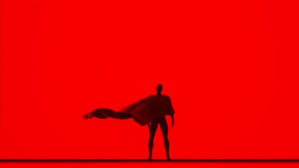 A photo of Superman standing with his back to a red background.