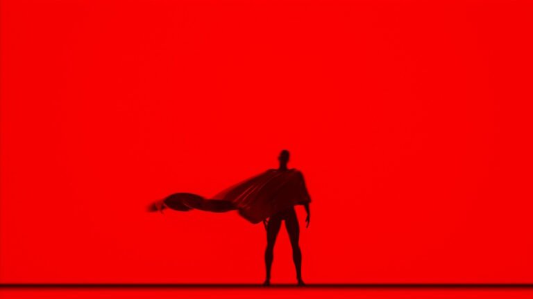 A photo of Superman standing with his back to a red background.