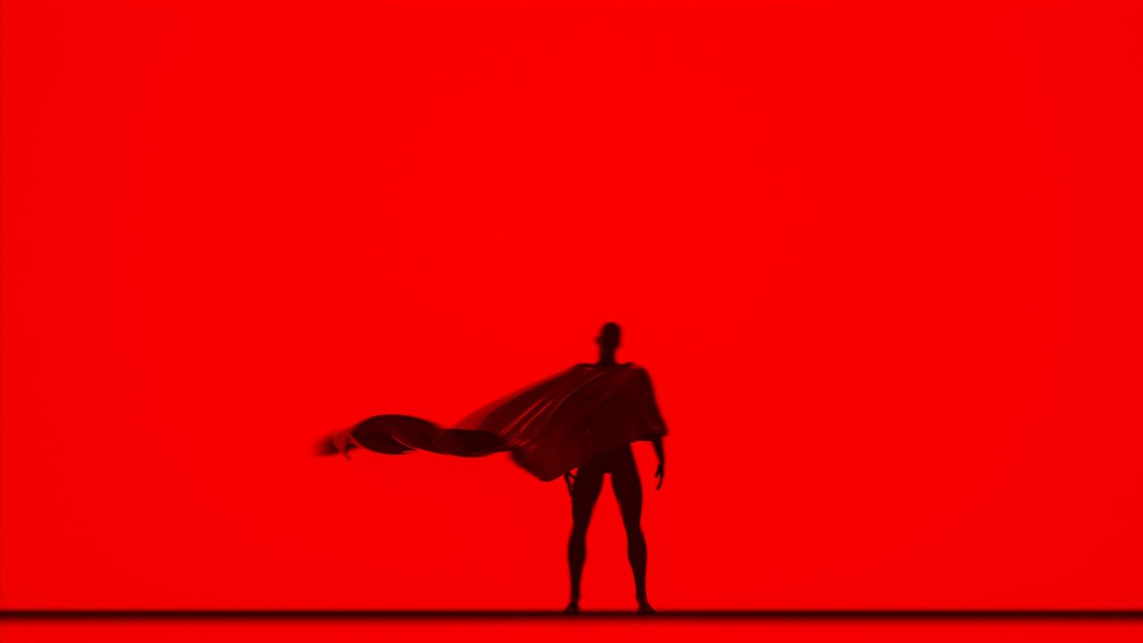 A photo of Superman standing with his back to a red background.